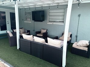 An outdoor seating area with wicker furniture and cushions, a flat-screen TV mounted on the wall, and a ceiling with exposed beams and string lights.