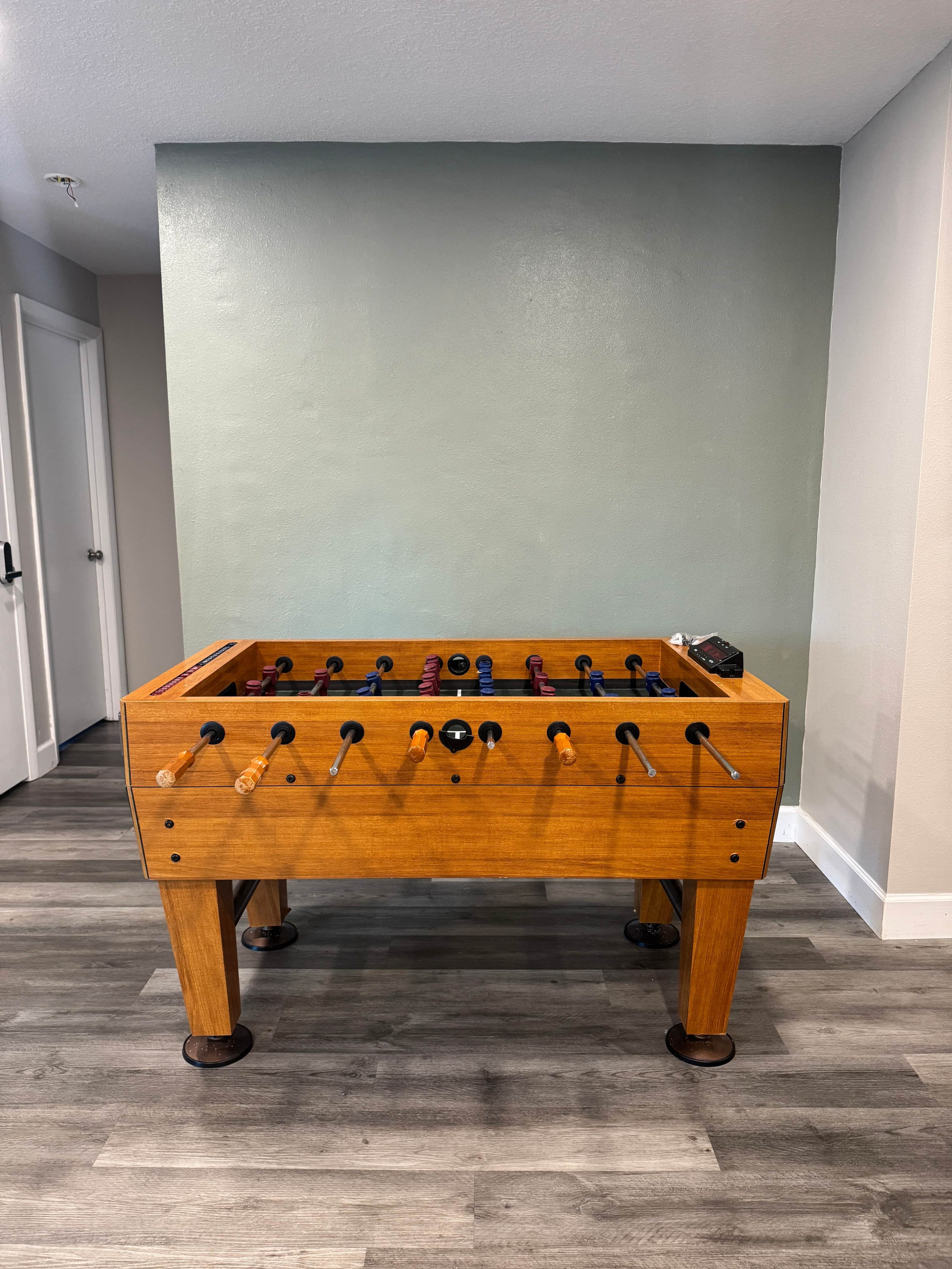 A wooden foosball table in a room with a wooden floor and light green walls.