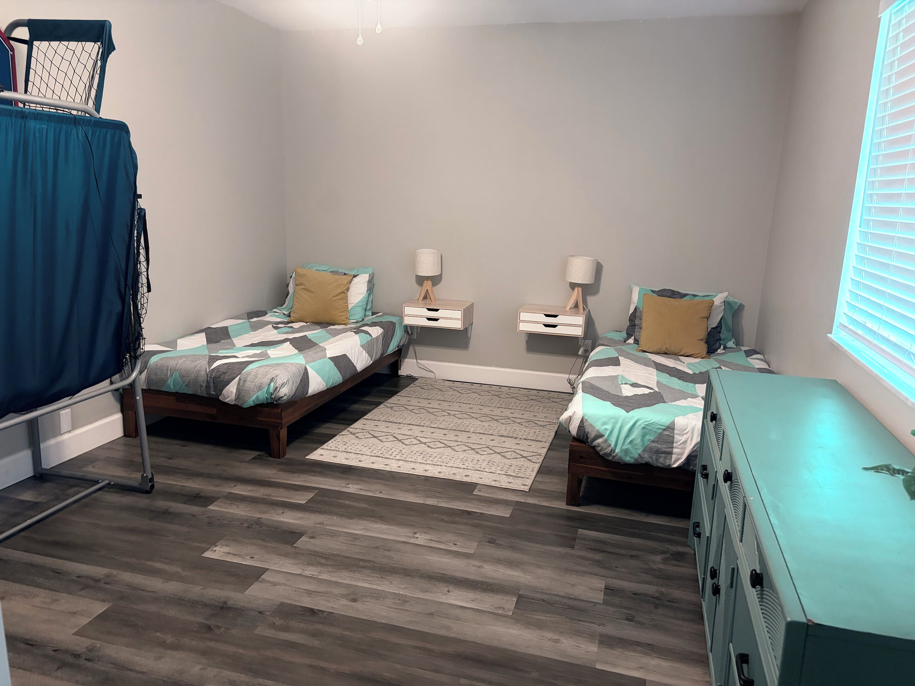 A bedroom with two beds, a small rug, bedside tables with lamps, a window with blinds, and a blue curtain.