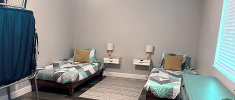 A bedroom with two beds, a small rug, bedside tables with lamps, a window with blinds, and a blue curtain.