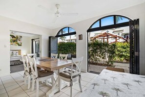 A dining area with a wooden table and white chairs, a ceiling fan, and a view of a garden through an open archway. - Mango Resort 2 Bedroom Pool Access 11 (Palm Cove)