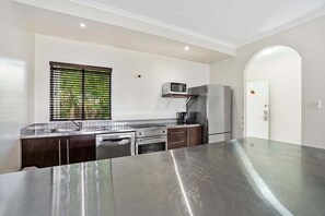 A modern kitchen with stainless steel appliances, a large island, and a window with blinds. - Mango Resort 11 | 2 Bedroom Pool Access Apartment (Palm Cove)