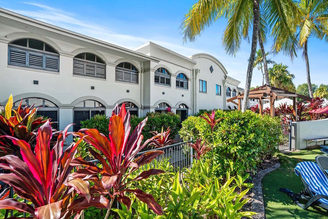 A hotel with a white facade, arched windows, and a well-maintained garden with tropical plants.