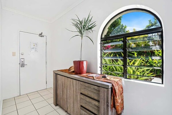 A room with a white door, a wooden cabinet, a potted plant, and a window with a view of greenery. - Mango Resort 2 Bedroom Pool Access 11 (Palm Cove)