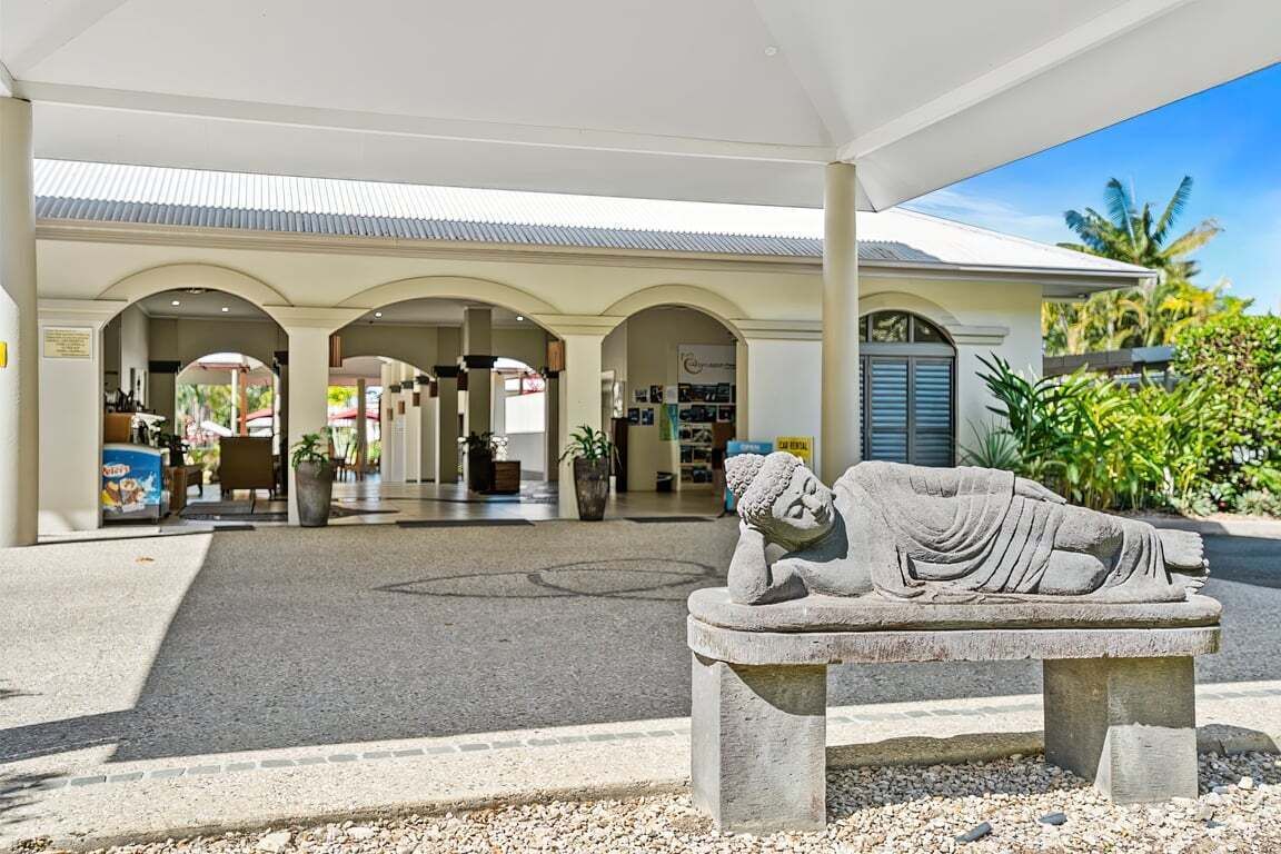 A stone statue of a reclining figure in a courtyard with a covered walkway and a building in the background.