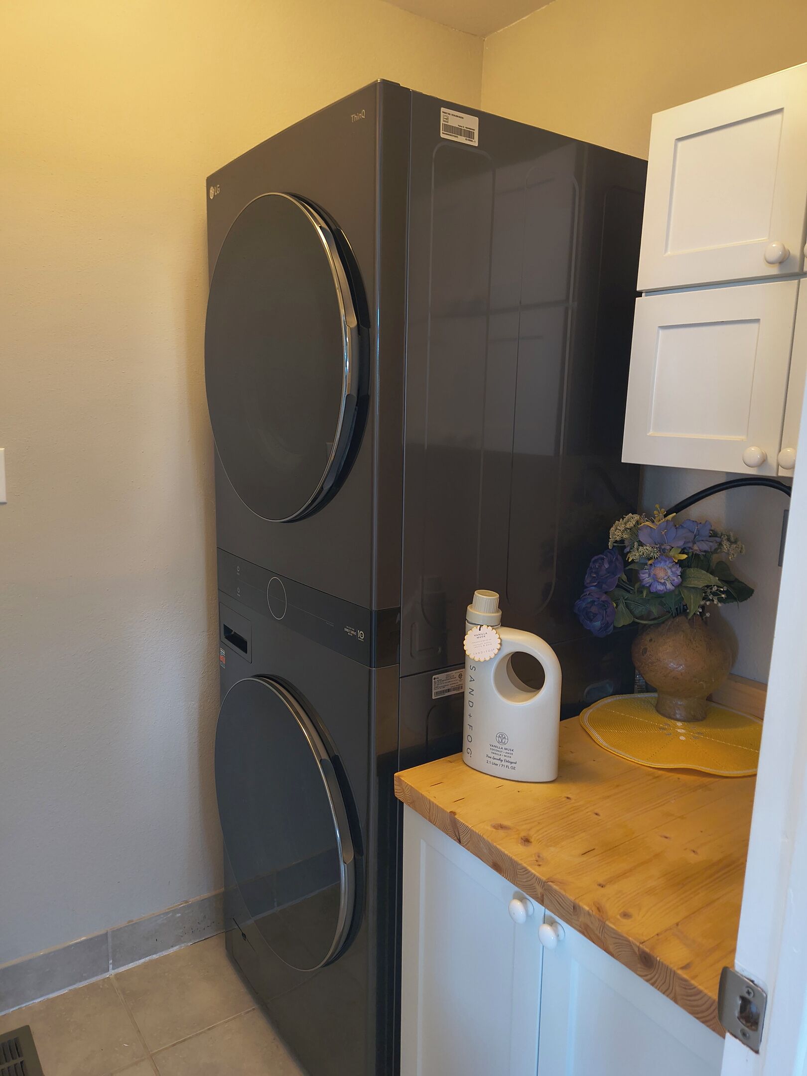 A compact laundry room with a front-loading washer and dryer, a small wooden counter with a bottle, and a white cabinet.