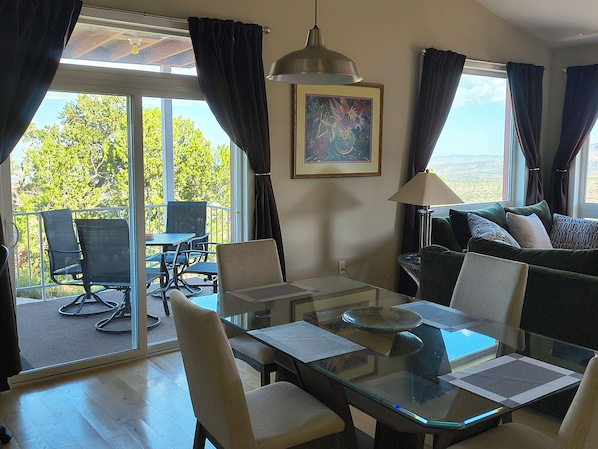 A room with a glass table, chairs, a sofa, a painting on the wall, and a view of greenery outside.