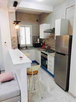 A compact kitchen with a central island, a built-in oven, a refrigerator, and a hanging light fixture. - The Wonderful “Sunset Bay” of Banlieue Nord (Zayatine)