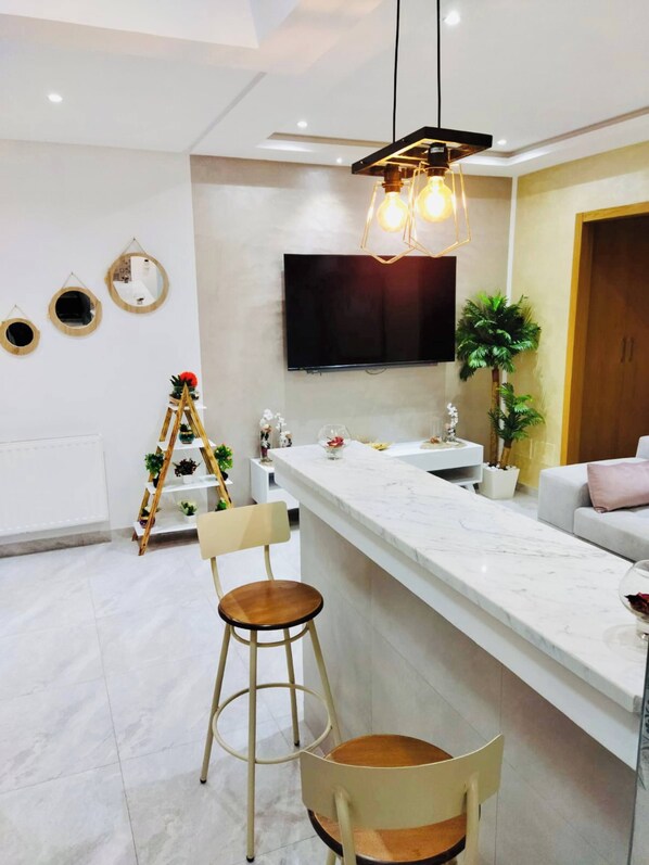 A modern kitchen with a marble countertop, a wooden chair, and a TV mounted on the wall. - The Wonderful “Sunset Bay” of Banlieue Nord (Zayatine)