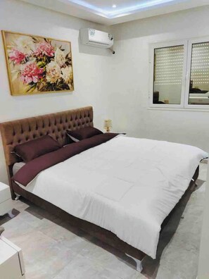A bedroom with a large bed, a wall-mounted air conditioner, a window with blinds, and a floral painting. - The Wonderful “Sunset Bay” of Banlieue Nord (Zayatine)