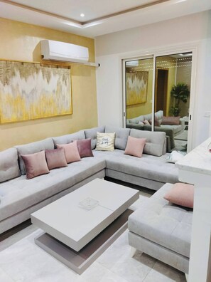 A modern living room with a grey sectional sofa, a white coffee table, and a large abstract painting on the wall. - The Wonderful “Sunset Bay” of Banlieue Nord (Zayatine)