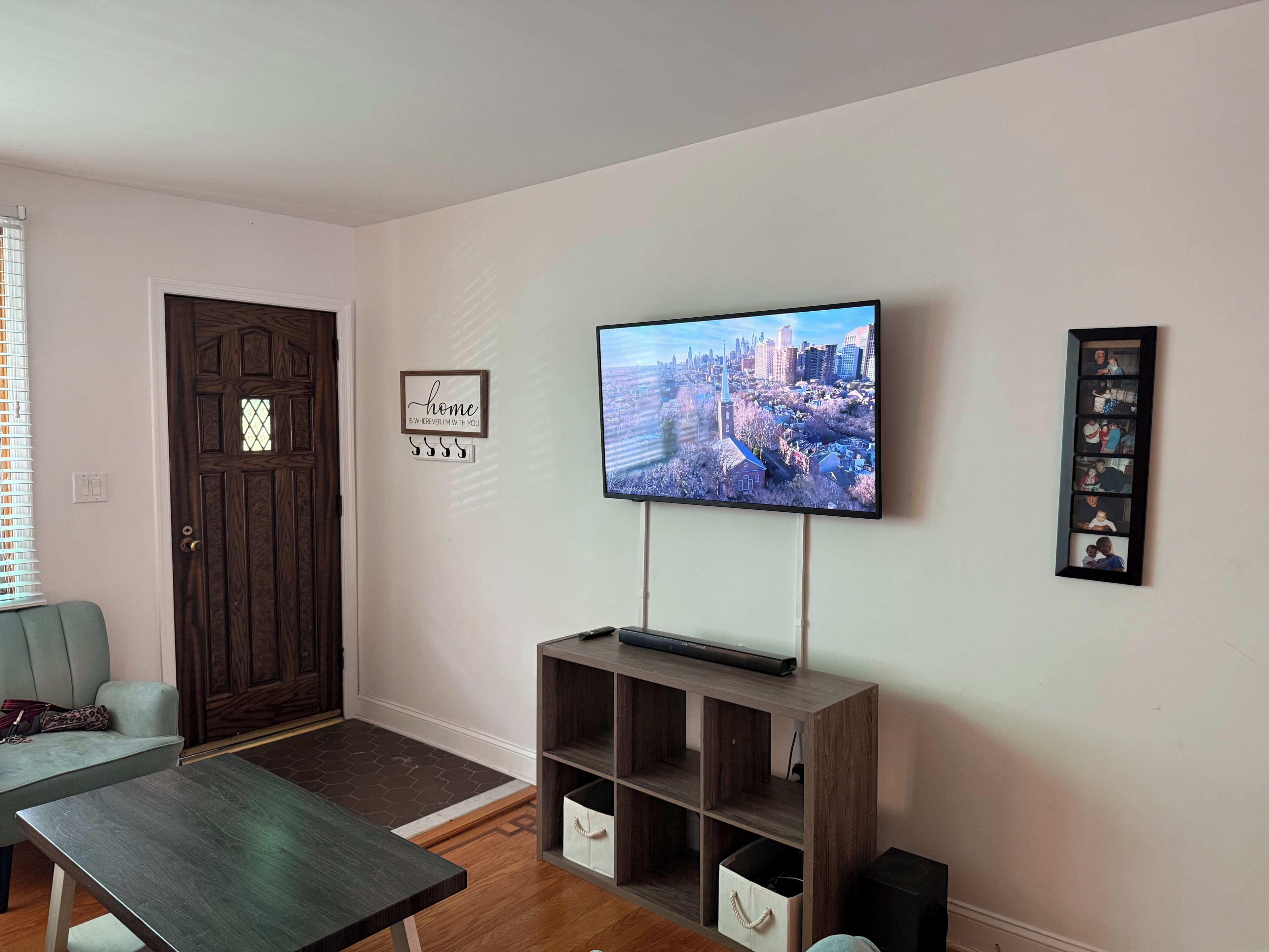 A living room with a flat-screen TV mounted on the wall, a wooden entertainment unit below it, a door with a decorative panel, and a framed quote on the wall.