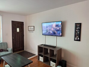 A living room with a flat-screen TV mounted on the wall, a wooden entertainment unit below it, a door with a decorative panel, and a framed quote on the wall.