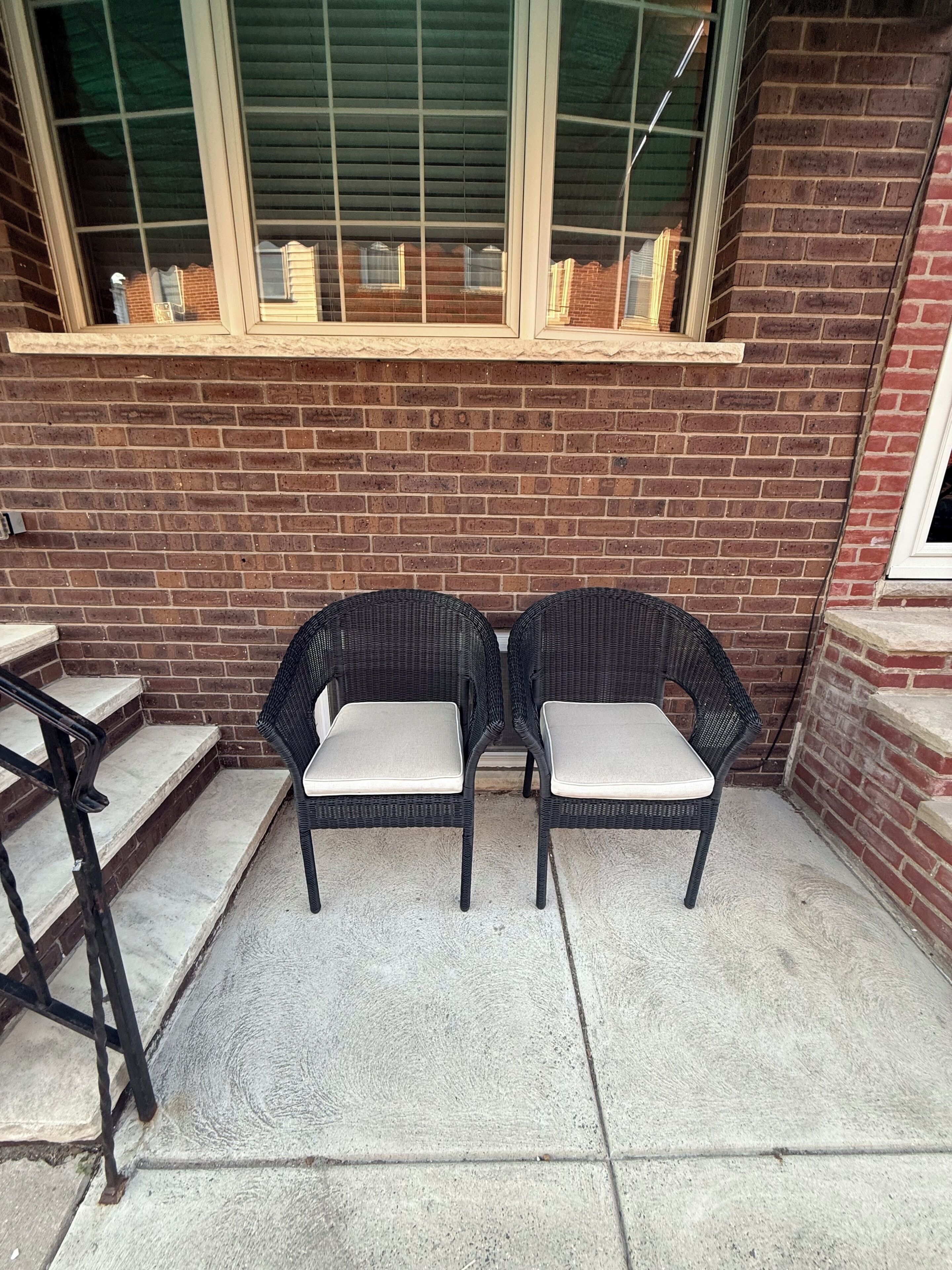 Deux fauteuils en osier, dotés d’assises et de dossiers rembourrés, sont placés sur une terrasse en béton, juste à côté d’un mur de briques surmonté d’une fenêtre.
