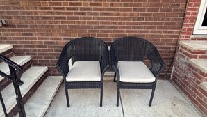 Two wicker chairs with cushioned seats and backs, positioned on a concrete patio adjacent to a brick wall with a window above.