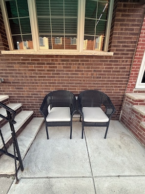 Two wicker chairs with cushioned seats and backs, positioned on a concrete patio adjacent to a brick wall with a window above.