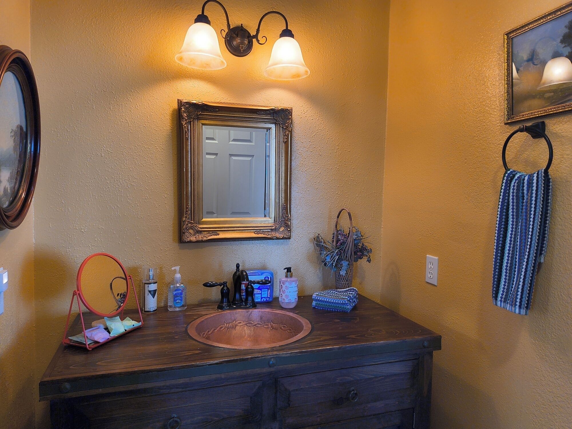 A bathroom with a wooden vanity, a mirror, wall-mounted lights, a towel rack, and a framed picture.