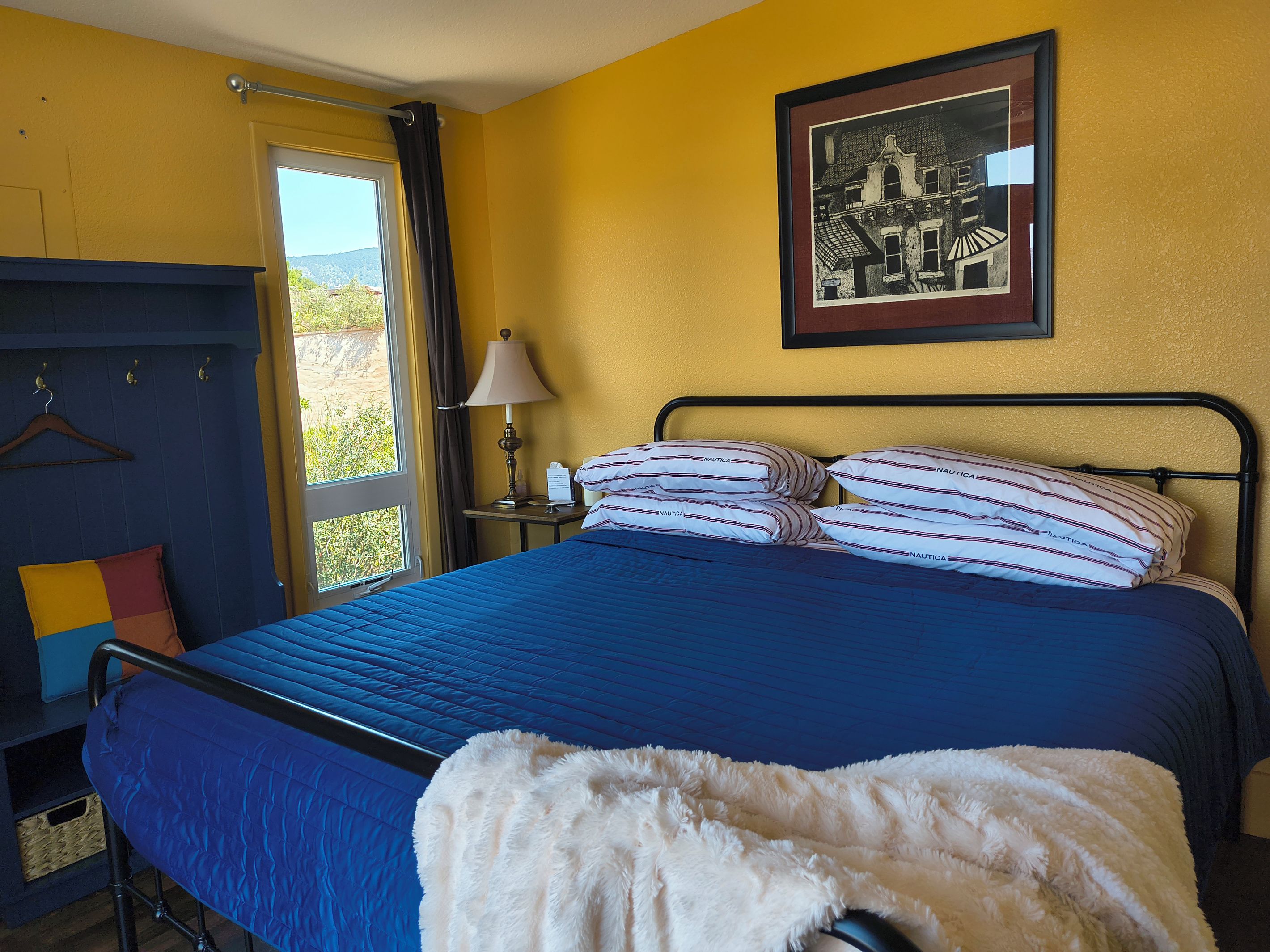A hotel room with a bed, a nightstand, a lamp, a framed picture on the wall, and a view of the outdoors.