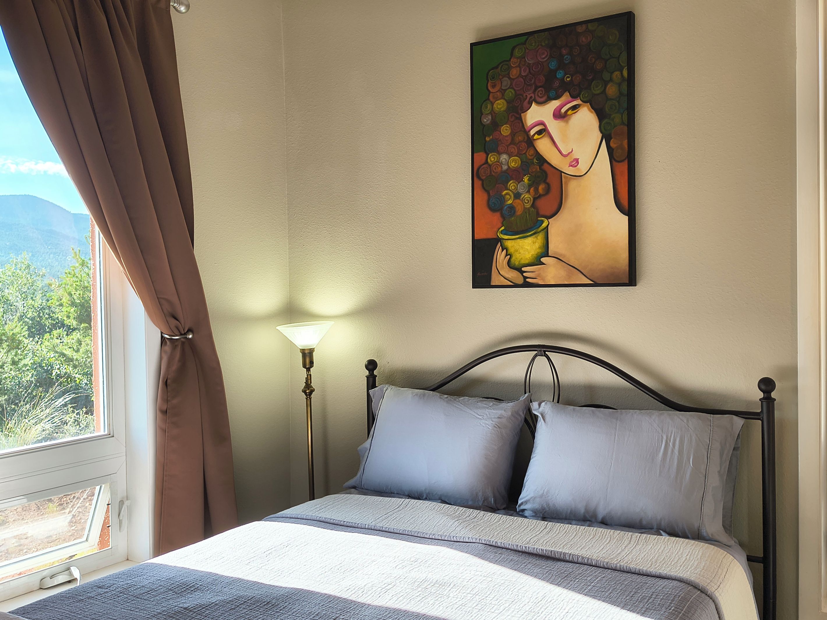 A bedroom with a bed, a painting on the wall, a lamp, and a window with curtains.
