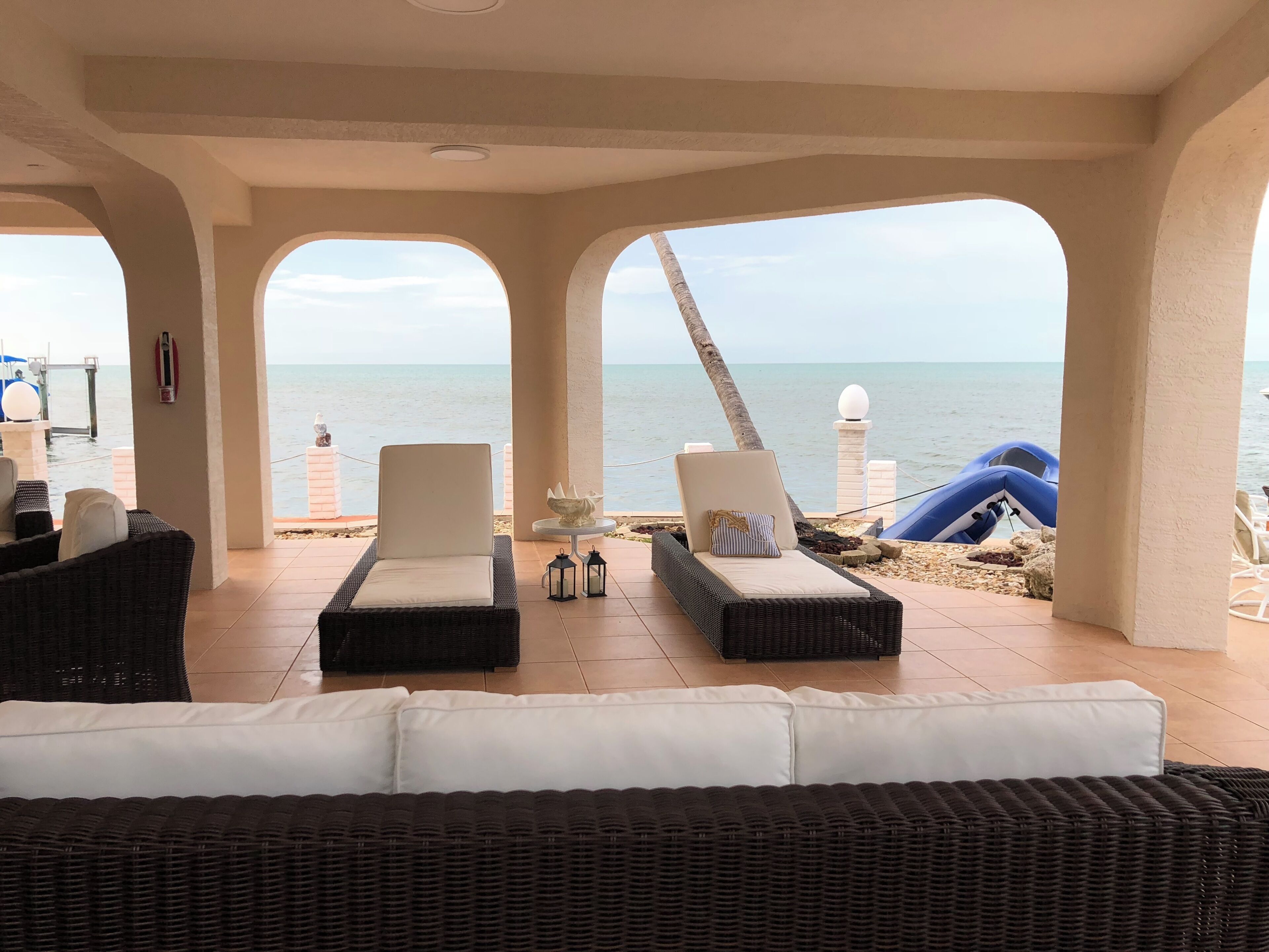 A covered outdoor seating area with lounge chairs and a sofa, overlooking a beach with a clear view of the sea.