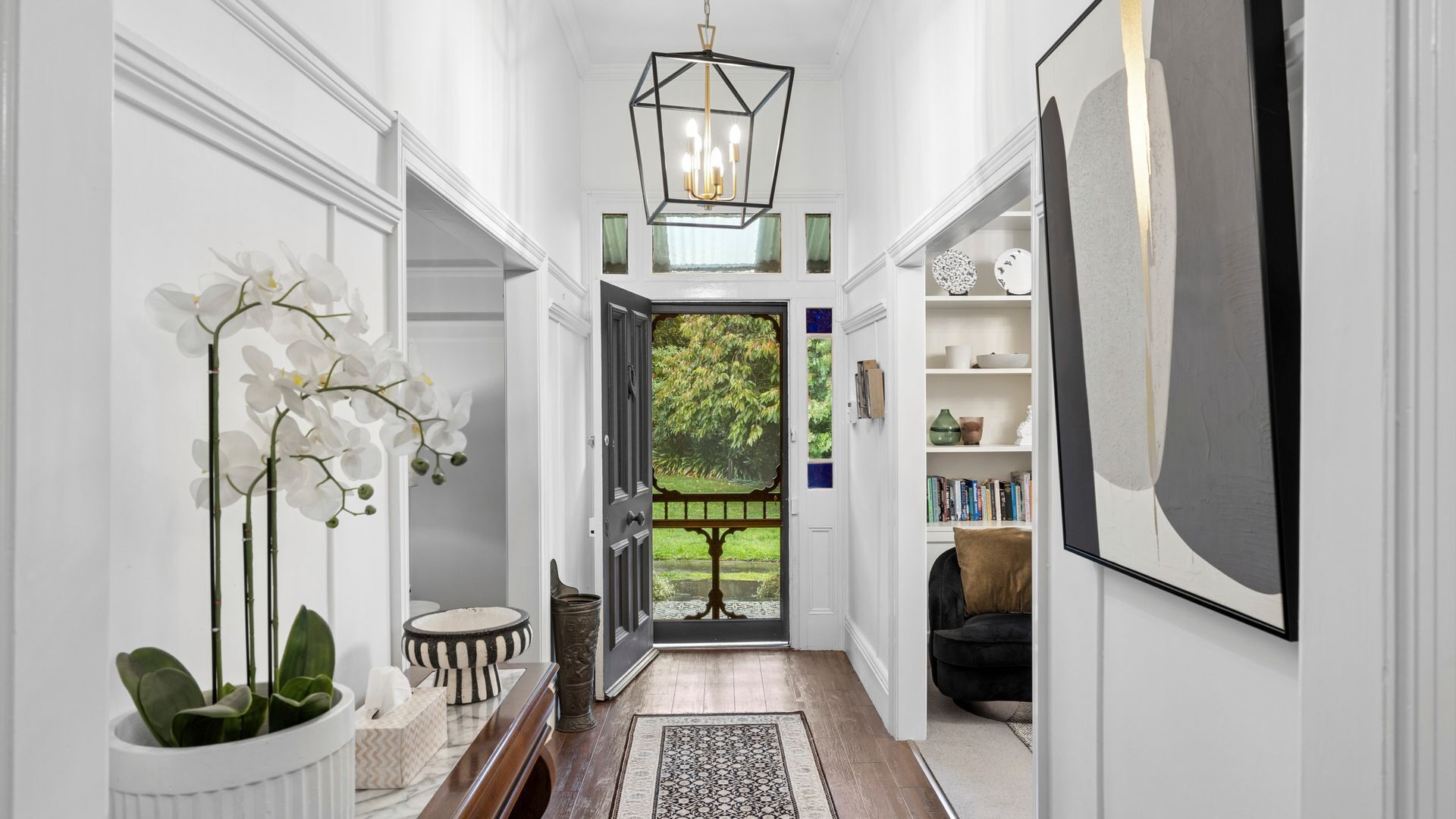A hallway with a wooden floor, white walls, a black and white patterned rug, a white potted plant, and a door leading to a garden.