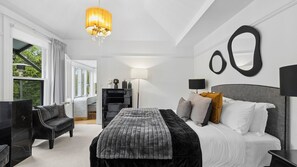 Ein modernes Schlafzimmer mit einem großen Bett, einem Schreibtisch und einem Sessel. Eine Pendelleuchte, eine Lampe und Blick ins Grüne sind vorhanden.