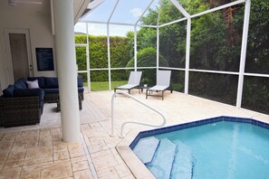 Área de piscina com estrutura de vidro, espreguiçadeiras e espaço de estar com um sofá.