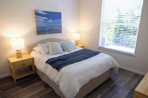 Quarto com cama, duas mesas de cabeceira, abajur, janela com persianas e um quadro na parede.
