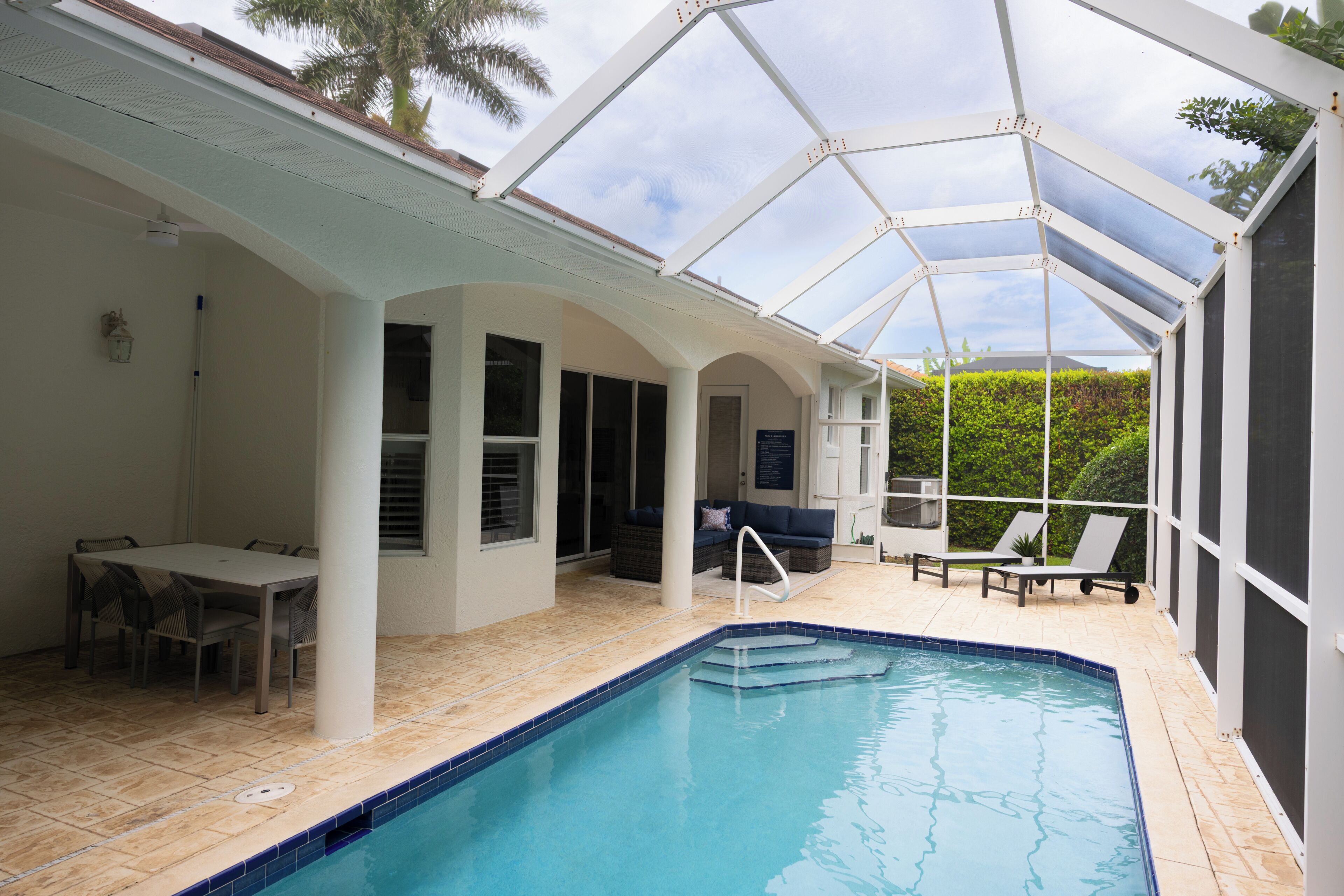 A covered outdoor swimming pool area with a dining table and chairs, a sofa, and lounge chairs.