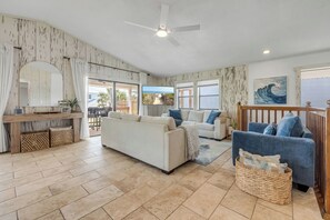A spacious living room with a large sofa, a blue armchair, and a wooden coffee table. There is a ceiling fan, a large window with a view of the outdoors, and a painting on the wall. - Bethune Beach 4BR Home with Rooftop Deck Walk to Beach and Indian River (New Smyrna Beach)