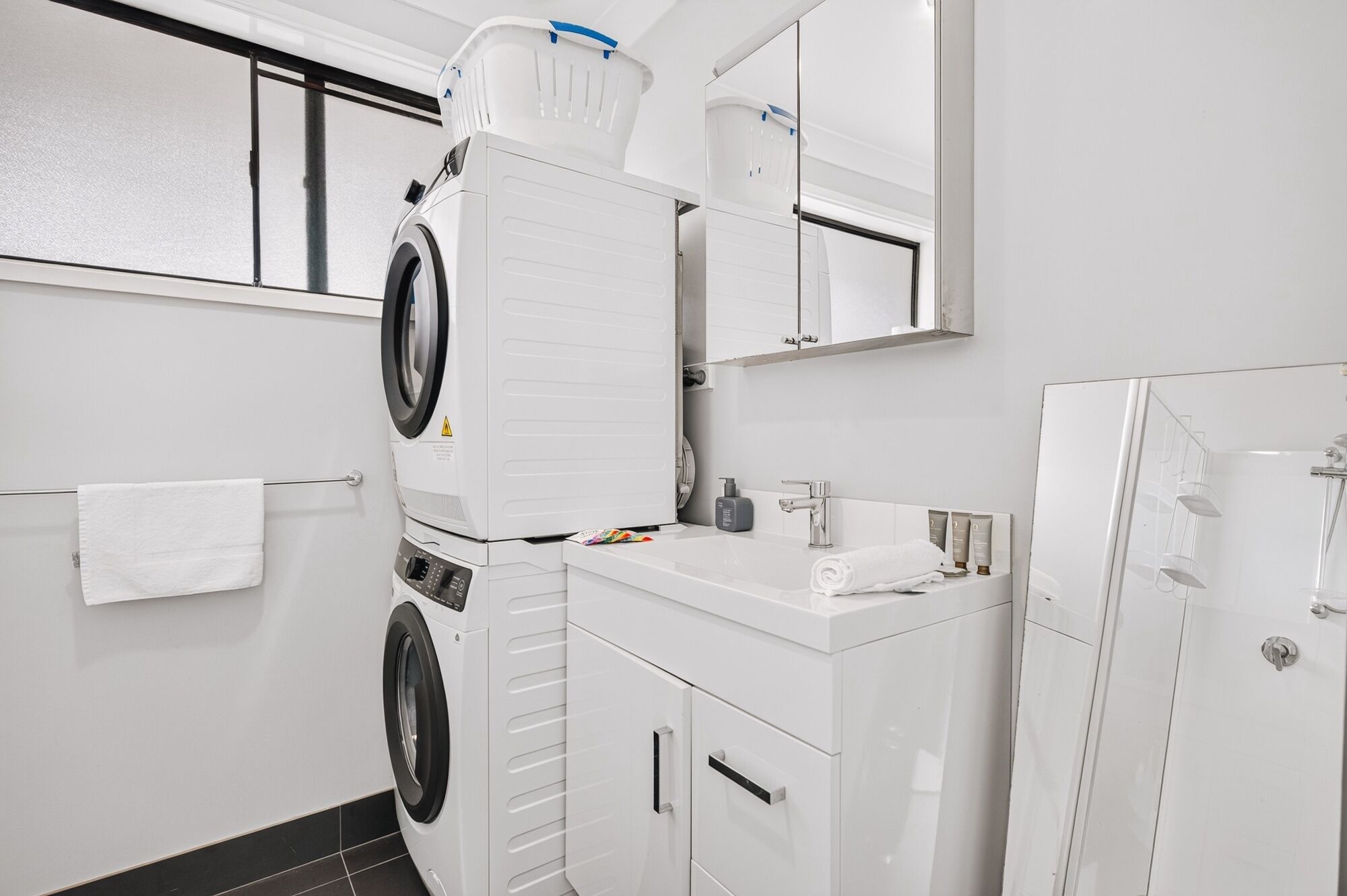 A modern bathroom with a stacked washer and dryer, a sink with toiletries, and a glass shower enclosure.