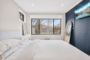 Une chambre avec un grand lit, une fenêtre donnant sur les arbres et une porte dotée d’un miroir.