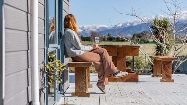 Une personne est assise sur un banc en bois devant un bâtiment, avec une vue sur les montagnes en arrière-plan.