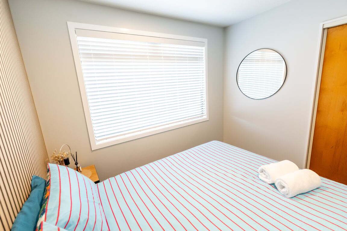 A bedroom with a bed, a window with blinds, a round mirror, and a wooden door.