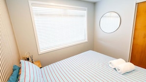 A bedroom with a bed, a window with blinds, a round mirror, and a wooden door.