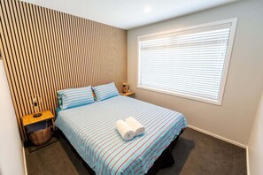 Hotelkamer met een bed, nachtkastje, lamp, raam met zonwering en wand met verticale lamellen.