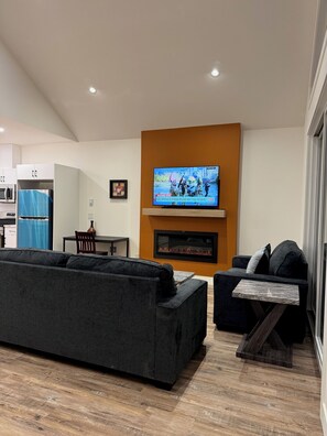 A modern living room with a large flat-screen TV mounted on a wall, a dark grey sofa, a smaller black sofa, and a wooden coffee table.