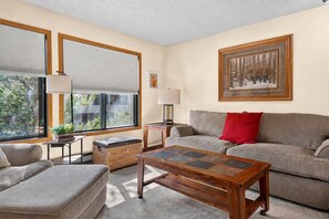 Ein Wohnzimmer mit einer Couch, einem Holztisch, einem Beistelltisch mit einer Lampe und einem Fenster mit Jalousien.