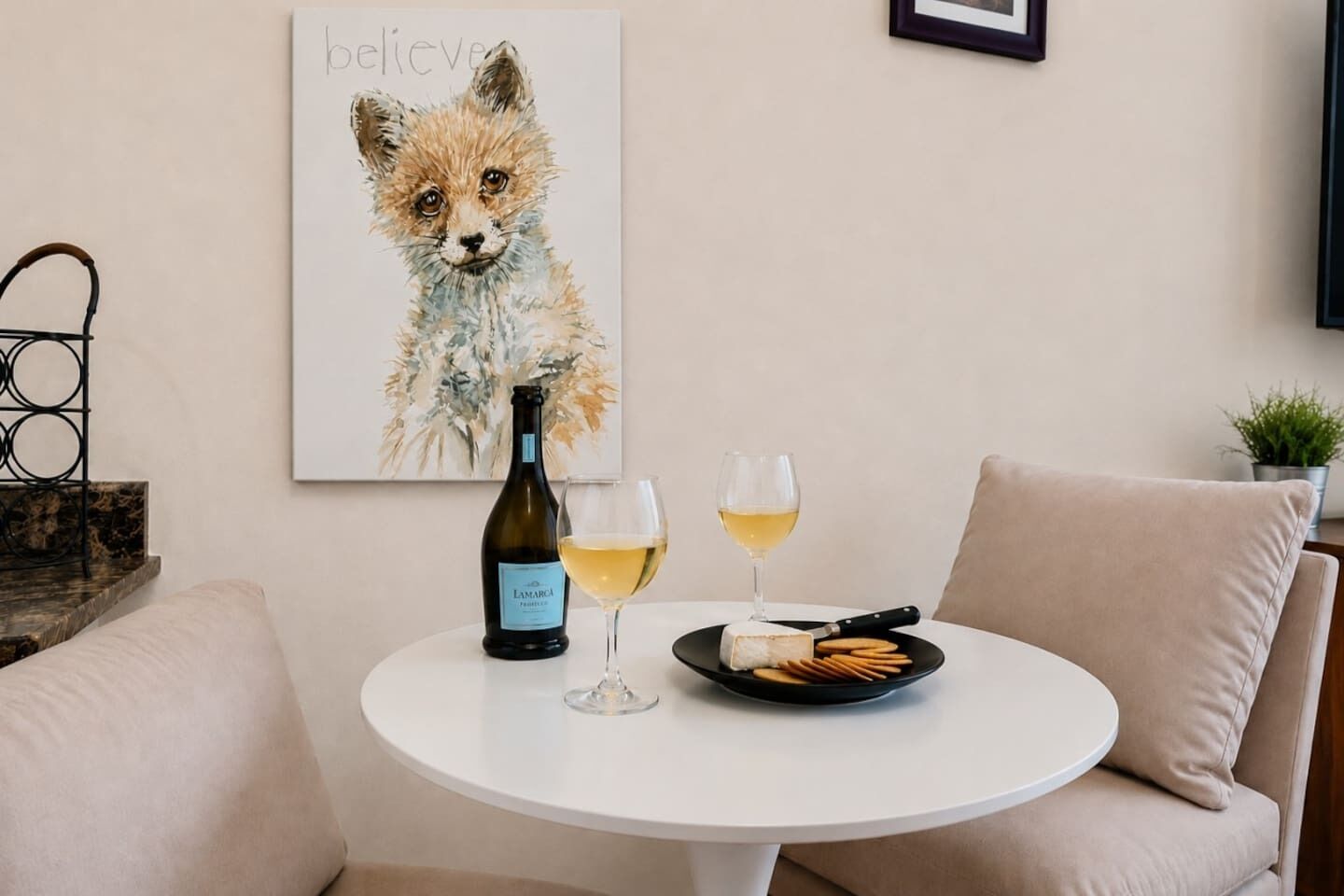 Ein runder weißer Tisch mit einer Weinflasche, zwei Gläsern, einer Platte mit Cräckern und einem Bilderrahmen mit einem Fuchs an der Wand.