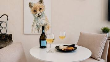 Ein runder weißer Tisch mit einer Weinflasche, zwei Gläsern, einer Platte mit Cräckern und einem Bilderrahmen mit einem Fuchs an der Wand.