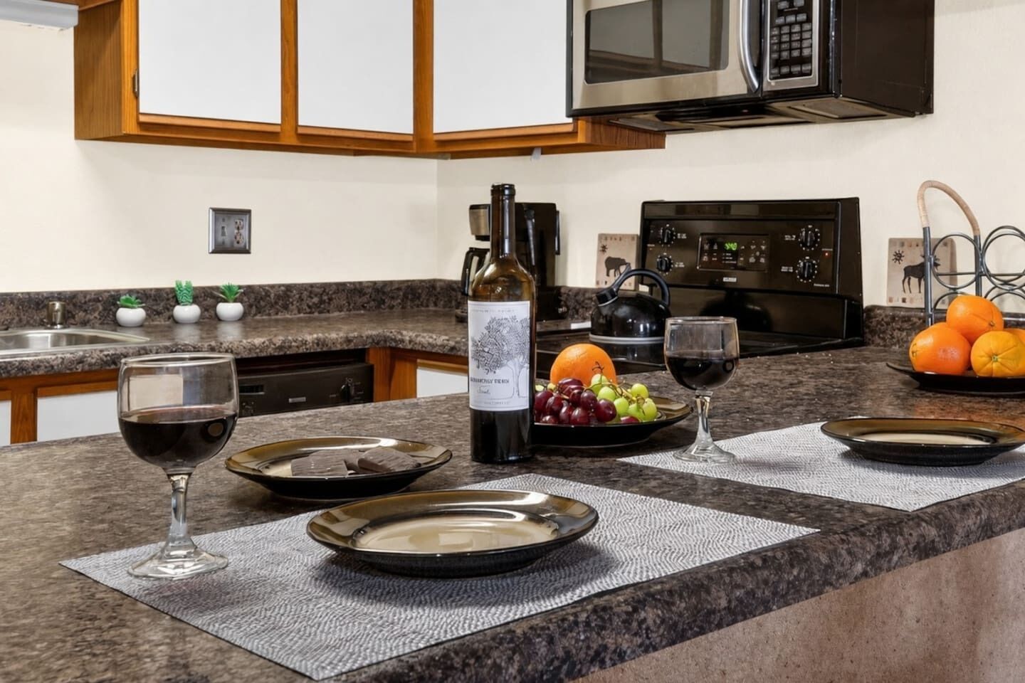 Une cuisine dotée de plans de travail en granit, d’une bouteille de vin, d’un verre de vin rouge, d’un panier de fruits et d’une bouilloire.