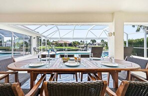 An outdoor dining area with a view of a swimming pool and a tropical landscape. - Baju | Gulf Access | Pool & Spa |  Game Room (Cape Coral)
