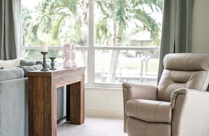 A living room with a sofa, a wooden side table, a fireplace, and a window with a view of palm trees. - Baju | Gulf Access | Pool & Spa |  Game Room (Cape Coral)
