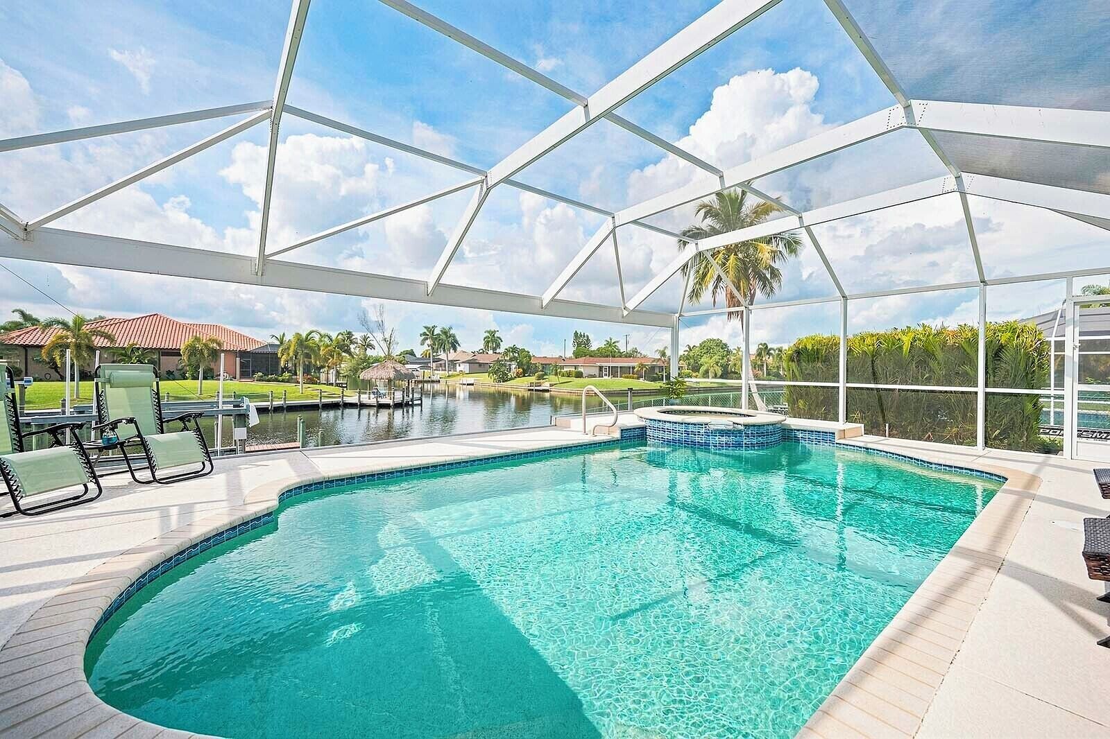 A pool area with a covered section, lounge chairs, and a view of a water body and palm trees.