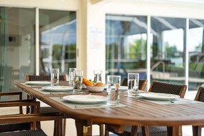 A dining table set with plates, glasses, and a bowl of fruit, with wicker chairs and a view of the outdoors through large windows. - Baju | Gulf Access | Pool & Spa |  Game Room (Cape Coral)