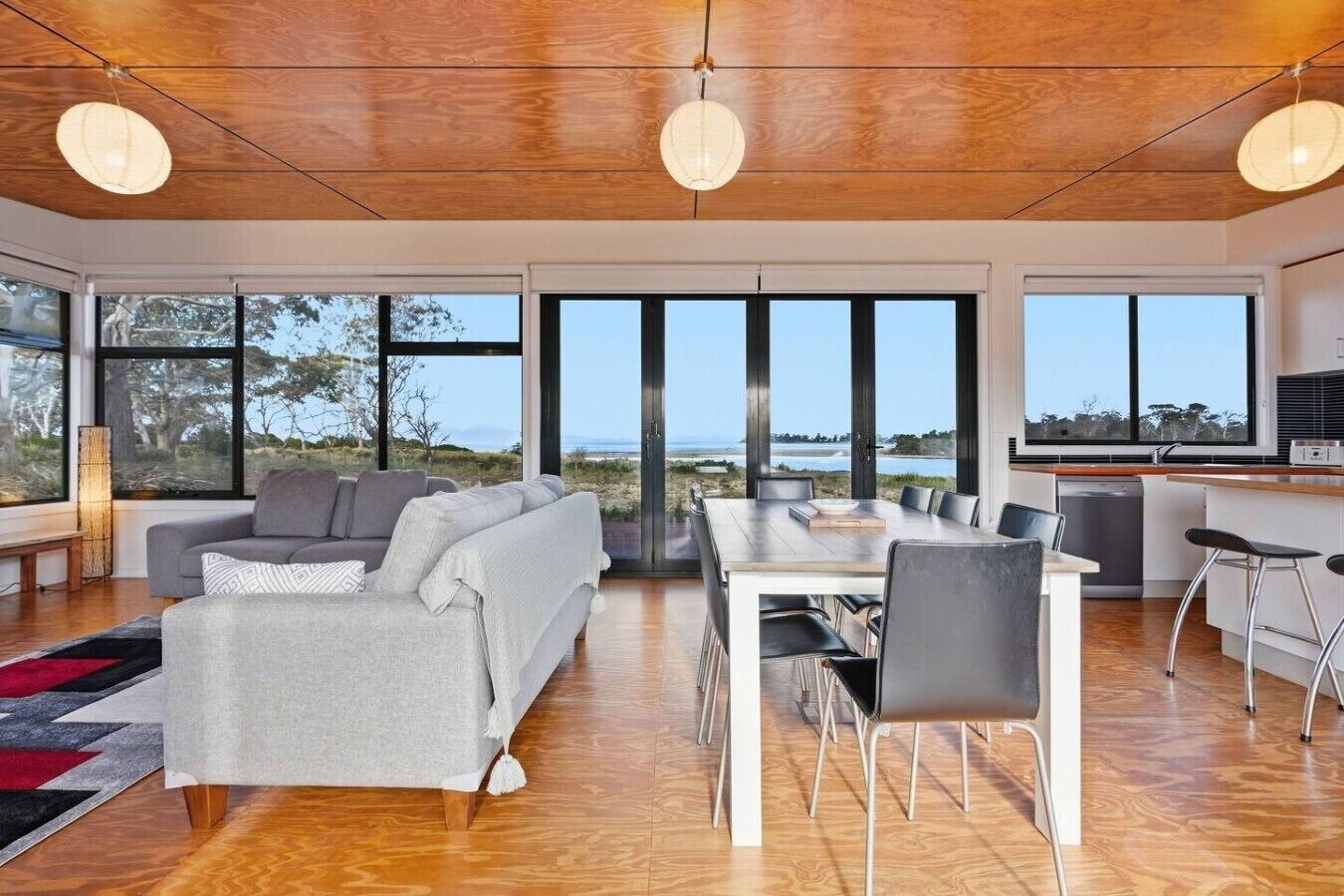 Ett modernt vardagsrum med en soffa, ett matbord och stolar, stora fönster med utsikt över havet och ett träpanelat tak.