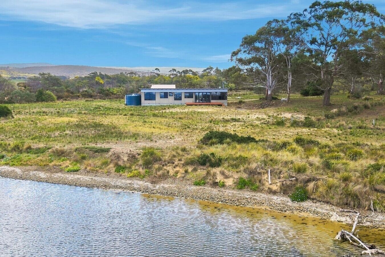 A modern house with a large glass window, situated on a grassy field near a body of water.