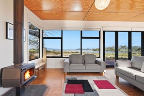 Un salon moderne avec un poêle à bois, un canapé gris et de grandes fenêtres donnant sur la plage.