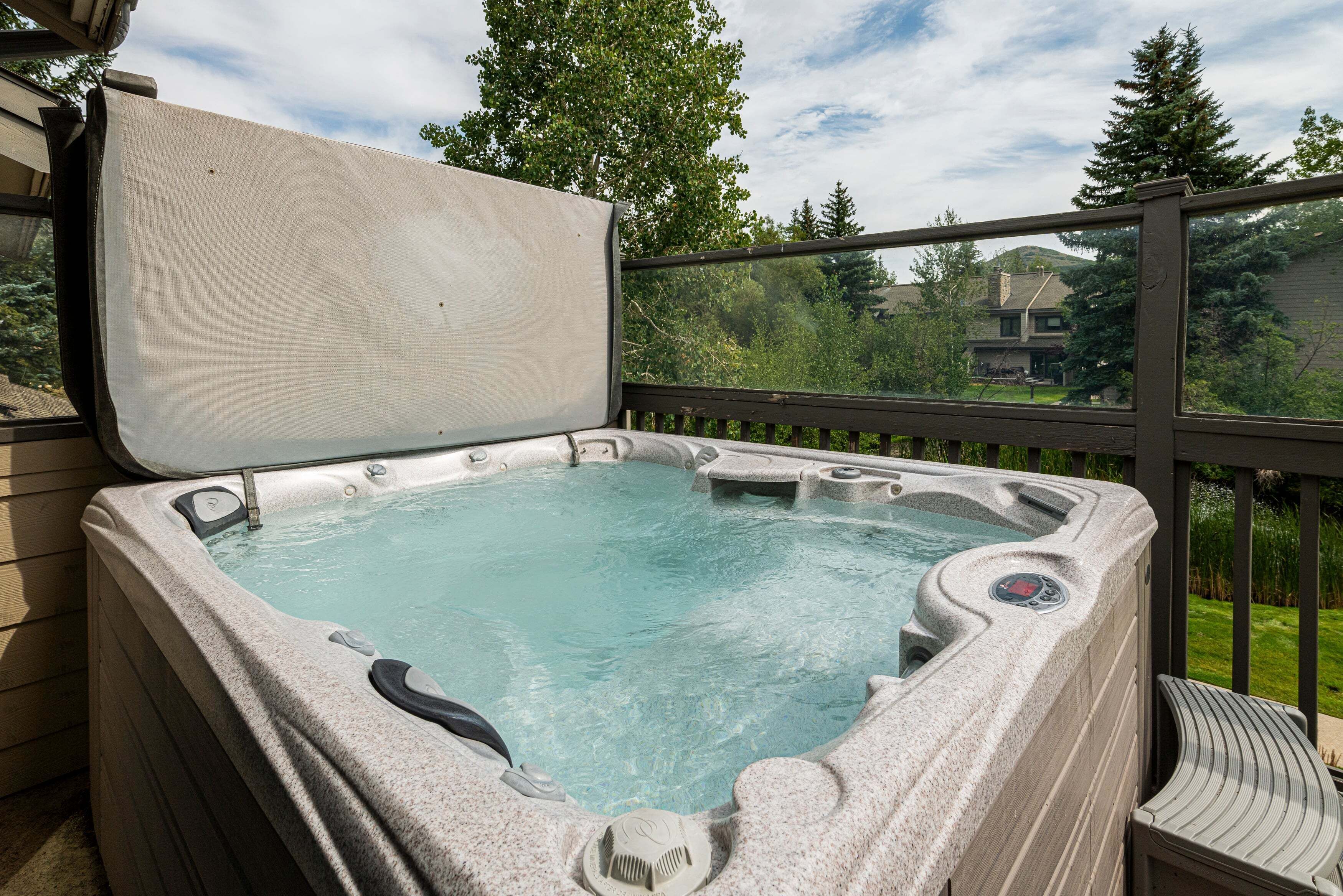 A hot tub with a cover, surrounded by a wooden deck and a glass railing, overlooking a residential area.