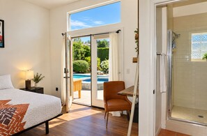 Une chambre à coucher avec un lit, une table de chevet, une lampe et une vue sur une piscine, visible à travers une porte ouverte.